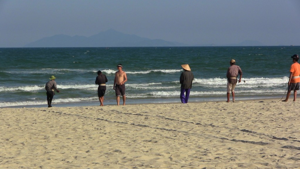 Vietnamese fishermen in Da Nang on November 30, 2013, pulling ashore their nets as a shirtless swimmer walks past them. (Credit: Nissa Rhee). 